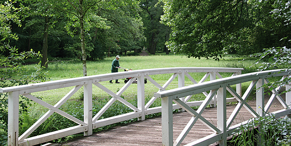 Blick von einer Brücke auf eine Wiese wo ein Gärtner den Rasen mäht.