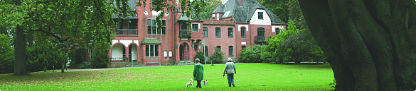 Rot geklinkertes Haus hinter Bäumen. Zwei Spaziergänger mit Hund gehen darauf zu. 